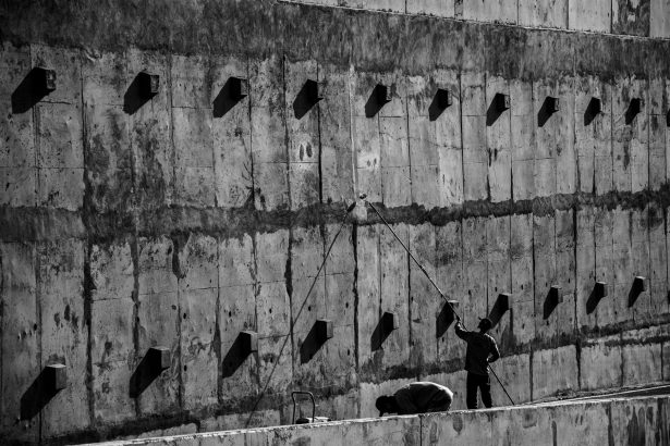 Dois trabalhadores pintam uma grande parede de concreto em um ambiente industrial.