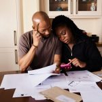 Casal organiza as finanças domésticas à mesa, analisando documentos e usando um smartphone.