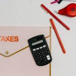 Calculadora, canetas e pasta com a inscrição “TAXES” sobre uma mesa de escritório.