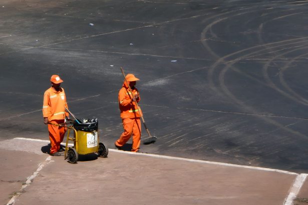 Dois trabalhadores da limpeza urbana atuam em uma rua de Brasília, usando uniformes laranja.